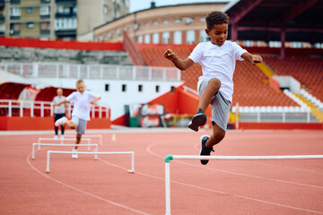Black little boy jumping over hurdles on running track at stadium.