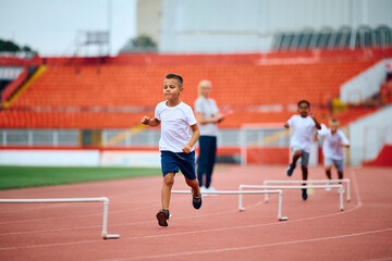 Obraz premium Little boy jumping over obstacles while running on track at athletics club.