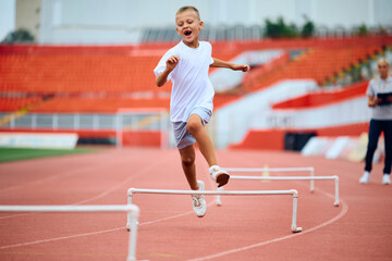 Fototapeta premium Cheerful kid umping over obstacles while running during sports training at stadium.