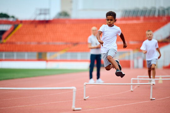 Black kid jumping over hurdles while running during exercise class at stadium.