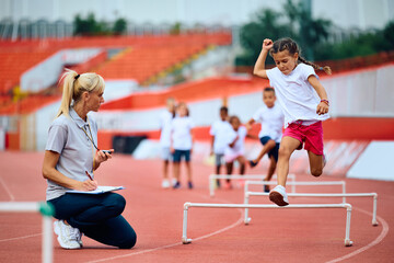 Little girl hurdling while PE teacher is measuring time on stopwatch at stadium.