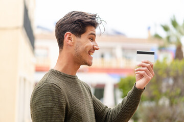 Young caucasian man holding a credit card at outdoors with happy expression