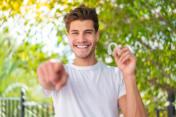 Young caucasian man holding invisible braces at outdoors points finger at you with a confident expression