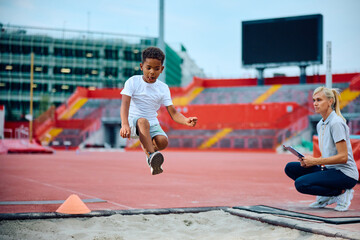 Black kid practicing long jump with his sports teacher at stadium. - obrazy, fototapety, plakaty