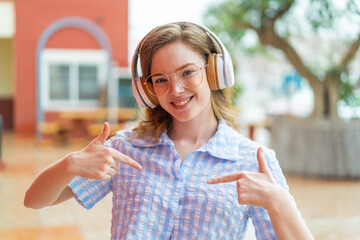 Young redhead girl headphones at outdoors proud and self-satisfied