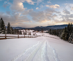 Secondary countryside alpine road in remote mountain village, snow drifts and wood fence on wayside