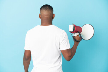 Young latin man isolated on blue background holding a megaphone and in back position