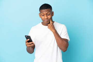 Young latin man isolated on blue background using mobile phone and thinking