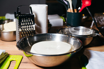 Dough Preparation in Stainless Steel Mixing Bowl. A stainless steel mixing bowl with dough for baking, next to a cheese grater and kitchen utensils on a wooden table.