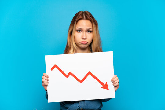 Teenager girl over isolated blue background holding a sign with a decreasing statistics arrow symbol with sad expression
