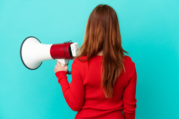 Teenager blonde girl over isolated blue background holding a megaphone and in back position