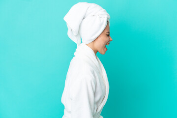 Teenager blonde girl in a bathrobe over isolated blue background laughing in lateral position