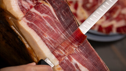 Top view of cutting acorn Iberian ham by knife with blur background with plate of pieces of Spanish...