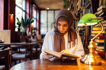 Portrait of serious Islamic teen girl in hijab reading book at library.