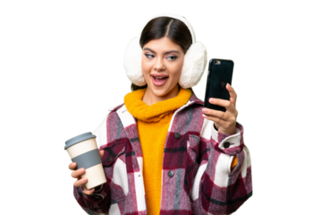 Young Russian woman wearing winter muffs over isolated chroma key background holding coffee to take away and a mobile