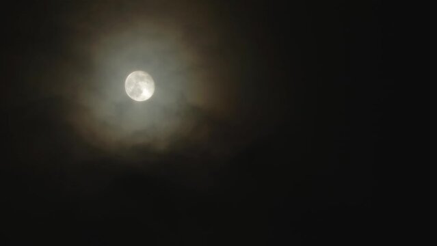 Full moon on a dark cloudy night with colored clouds flying in the sky in front of the Moon. Corona - light optical phenomenon