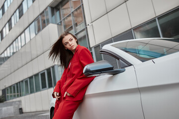 Pensive stylish young brunette in red trouser suit standing by convertible