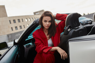 Stylish confident young brunette in red suit sitting in white convertible