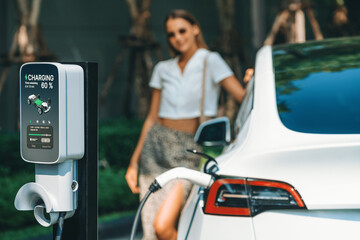 Young woman travel with EV electric car charging in green sustainable city outdoor garden in...
