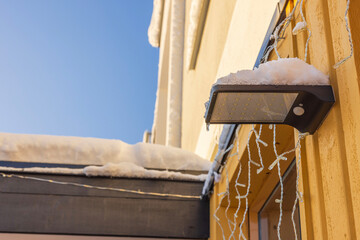 Detailed view of outdoor lamp featuring snow-clad solar panel on facade of villa on crisp winter...