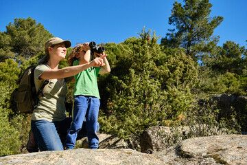 Naklejka na ściany i meble Smiling traveling young mother and little exploring forest with binoculars