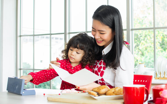 Beautiful Asian woman wearing apron and little cute daughter girl, giving gift and flowers on birthday or valentine day, smiling with happiness, having breakfast together,playing in kitchen at home.
