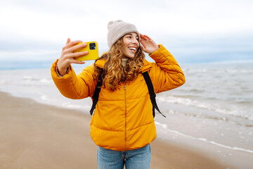 Portrait of a happy woman with a phone on the seashore. She is wearing a bright yellow jacket. Young woman taking a selfie and enjoying the scenery outdoors. Adventure concept, vacation.