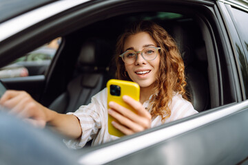 Portrait of a female driver uses a smartphone while driving a car. A young woman in a car uses a...