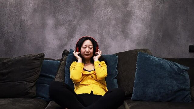 Young Asian Woman is Lying at Home on the Couch with her Eyes Closed and Listening to Music on Headphones. Woman is Resting and Hearing to Online Radio. Relaxation, Meditation and Mindfulness.