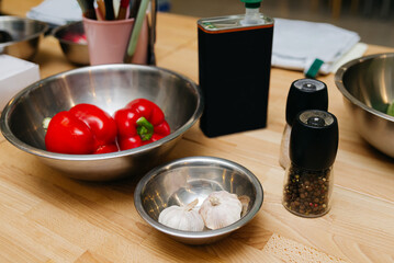 Kitchen Prep with Fresh Bell Peppers and Garlic. Red bell peppers and whole garlic cloves in stainless steel bowls with a pepper grinder, all set on a wooden countertop for meal preparation.