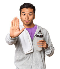 Chinese man with sportswear, towel, bottle in studio standing with outstretched hand showing stop...