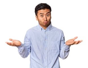 Young Chinese man in studio background doubting and shrugging shoulders in questioning gesture.