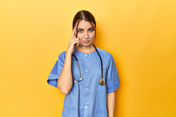 Young nurse in a yellow studio pointing temple with finger, thinking, focused on a task.