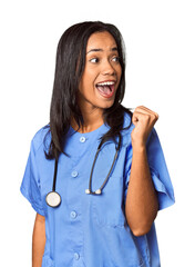 Young Filipina nurse posed in studio points with thumb finger away, laughing and carefree.