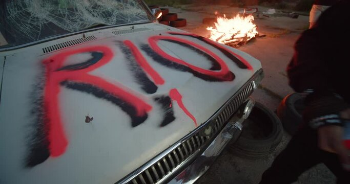 Riot Fire. Protest. Vandalism Punk protester girl uses a red pressure paint finishing vandal graffiti riot ugly lettering on viciously crushed old car on the street