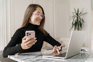 Smiling woman multitasking with a smartphone and laptop in a chic home office, dressed in a stylish...