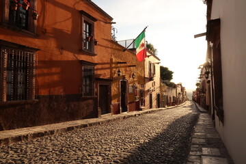 Typical cobblestone street in San Miguel de Allende at golden hour/sunset, a mexican flag is hanging on a facade, Mexico