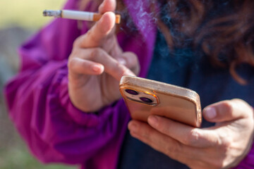 Woman's hand holding a smoking cigarette while using her mobile phone. Concept: vice