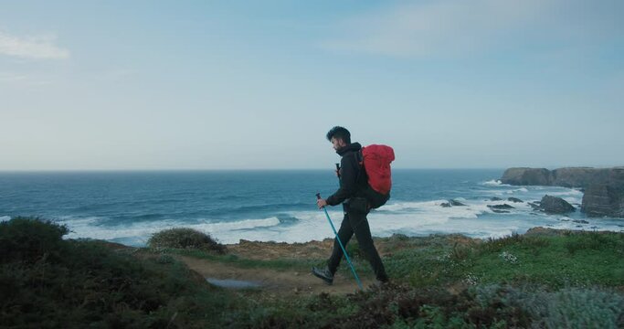 Side shot with gimball of hispanic young man walk on empty trail on cliffs along ocean. Windy hiking day in solitude. 