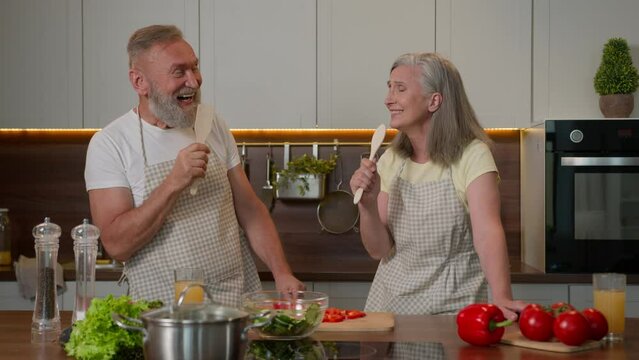 Funny Caucasian couple in love mature middle-aged senior old wife and husband retired married family singing song at spoons kitchenware joyful laughing woman man happy cooking healthy food in kitchen