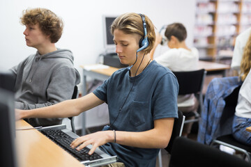 Portrait of a fifteen-year-old schoolboy in headphones, studying at a computer in the classroom at...