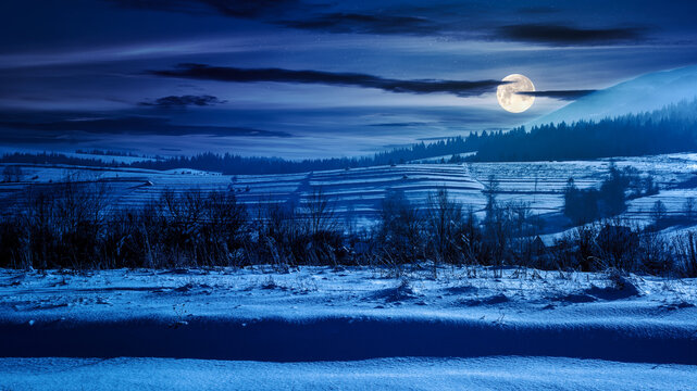 mountainous rural landscape on a winter night. countryside scenery with snow covered hill in full moon light