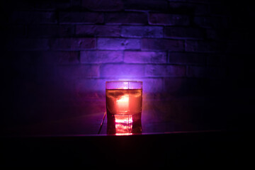 Whiskey in fire concept. Glass of whiskey and ice on wooden surface with color light and fog on...