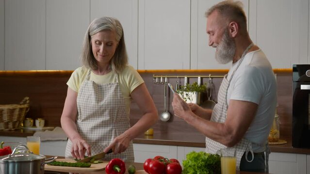 Caucasian couple middle-aged family woman man cook breakfast in home kitchen senior wife chop fresh vegetables healthy food mature husband take photo record video blog on mobile phone cooking dinner