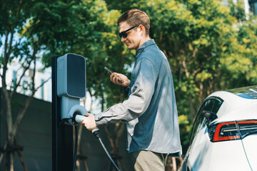 Young man travel with EV electric car charging in green sustainable city outdoor garden in summer...