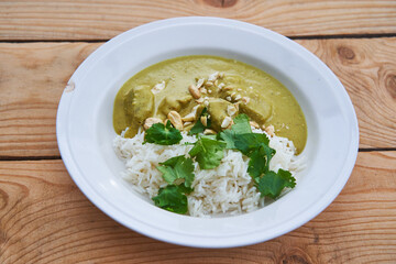 Sweet indian korma curry made from lamb meat, coconut milk, casheew nuts and lot of vegetables served with basmati rice and green coriander leaves in the deep rustic porceain plate on the wooden table