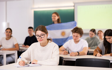 A young female student is carefully recording a lecture by a school teacher