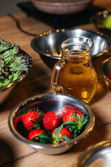 On a wooden table, a set of food ingredients, a pitcher of vegetable oil, and a steel bowl with strawberries