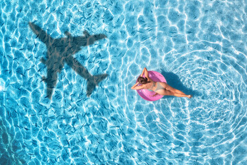 Aerial view of a young woman swimming with pink swim ring in blue sea with airplane shadow in water in summer. Tropical landscape with girl, plane shadow, clear water, sandy beach. Top view. Travel