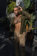 Chico joven musculado y tatuado posando con ropa urbana y moderna en zona de bares con palmeras al atardecer